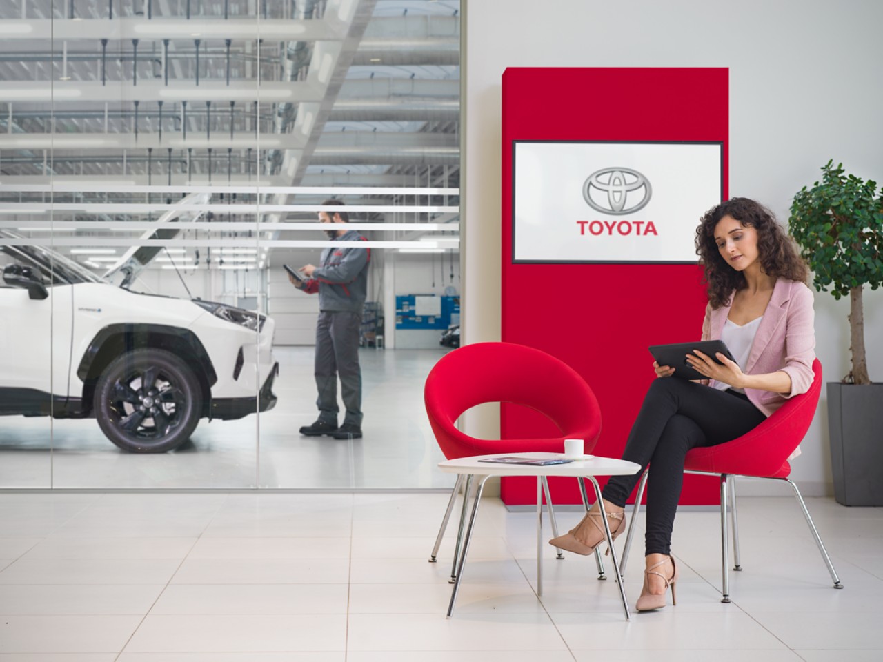 A women sits in a Toyota Service waiting area. She looks at a tablet, with a Toyota logo displayed on a tv screen behind her.